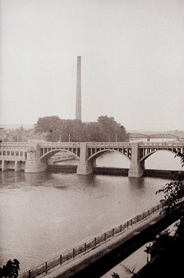 A concrete arch bridge spans a river, flanked by dense foliage and a prominent industrial chimney rising in the background. T...