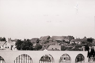 A wide view of rooftops in what appears to be a small town, possibly Eastern Europe. A man in a suit stands near a decorative...