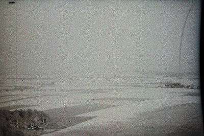 Monochromatic landscape shows snow-covered rolling hills and sparse vegetation. A vehicle is visible on a winding road, hinti...