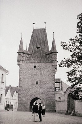 A stark, stone gateway tower with a slate roof and corner turrets dominates this view. Three figures are silhouetted walking ...