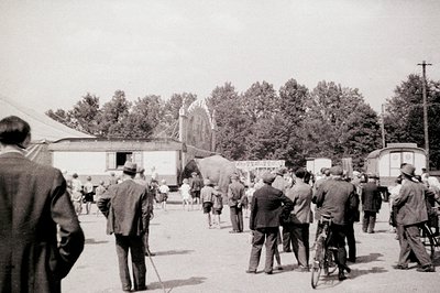 Group of formally dressed men & children gather near a large tent, potentially a fair or circus. Vintage attire suggests a mi...