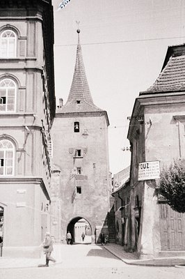 Stone gate tower with a conical roof, flanked by buildings with detailed facades. The street slopes downwards, showing a smal...