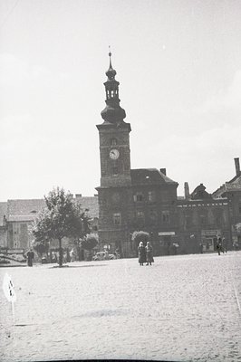 Grand, red-brick town hall with a clock tower dominates a wide, cobblestone square. Several figures populate the foreground, ...
