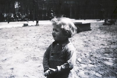 A young child with curly hair stands outdoors, gazing off-camera. They wear a sweater and overalls. The background shows tree...