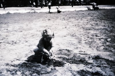 A young child crouches in the sand, engaged with a small pail and shovel. Shallow depth of field focuses on the child; blurre...