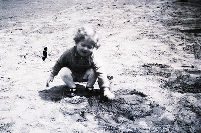 A young child, crouched in a shallow depression, appears to be playing in wet sand or soil. The child wears a short-sleeved o...