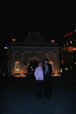 Two young individuals stand before an ornate stone gateway, likely an entrance to a park or grand building. The architecture ...