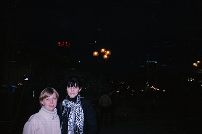 Two young women stand outdoors at night. One wears a white sweater, the other a dark coat with a leopard-print scarf. Backgro...