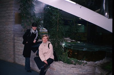 Two women pose in a large, indoor space featuring a rock waterfall and koi pond. The architecture includes a curving white st...
