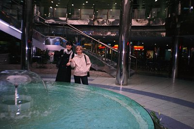A wide-angle view captures two women standing beside a circular fountain within a multi-level shopping complex. Modern archit...