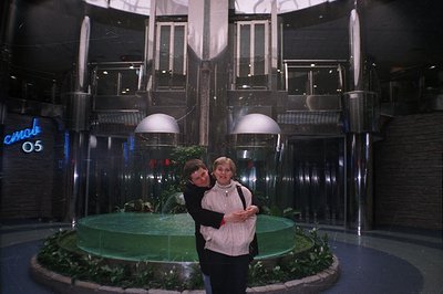 A couple stands embracing near a tiered fountain inside a large, modernist building, likely a shopping center. Visible signag...