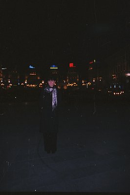 A young woman stands outdoors amidst falling rain, a backdrop of blurred, warmly lit buildings visible. She wears a dark coat...