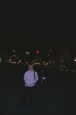 A young man stands in a city street at night, facing the camera. He wears a light-colored, textured jacket over dark pants, a...