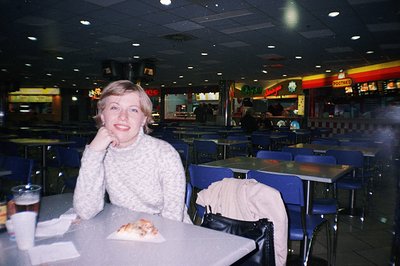 A young woman sits at a table in a bustling food court, likely a shopping mall. She wears a dark sweater, with a slice of piz...