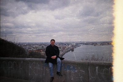 A man in a dark coat sits casually on a concrete ledge overlooking a cityscape & river. Notable film grain & light leaks indi...