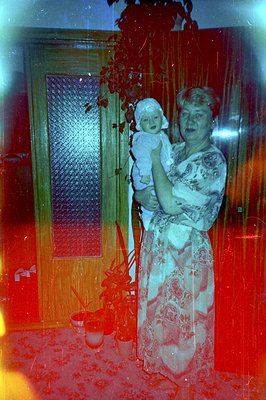 A woman in a patterned dress cradles an infant in a brightly lit interior. Potted plants flank a partitioned doorway. The ima...
