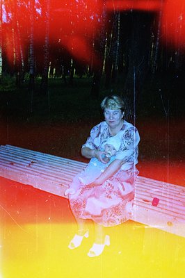 A woman sits on a textured wooden bench, holding a baby swaddled in pink. Background of tall, dark trees suggests a park or w...