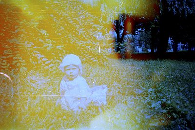 A young child, likely an infant, kneels in tall grass surrounded by foliage. Noticeable color shift and light leaks indicate ...