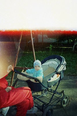 A baby sits in a vintage, bulky stroller featuring a striped padded interior. The child wears a light blue outfit & white cap...