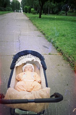 Infant rests in a vintage, dark green baby carriage, covered with a patterned cloth. The carriage sits on a cracked, wet conc...