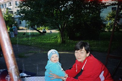 A candid snapshot of a mother and young child in a park setting. The child, wearing a light blue hooded towel and matching ha...