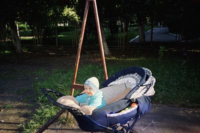 A baby sits in a dark blue stroller with a patterned blanket, wearing a light blue outfit and white hat. Background shows a m...