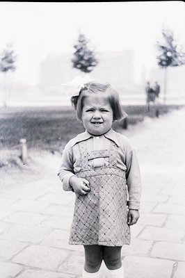 A young girl stands outdoors, wearing a plaid dress with suspenders and white socks. A large ribbon adorns her short hair. Th...