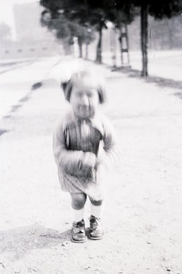 A young child, possibly a toddler, stands facing the camera with a slightly pensive expression. They wear a short-sleeved dre...