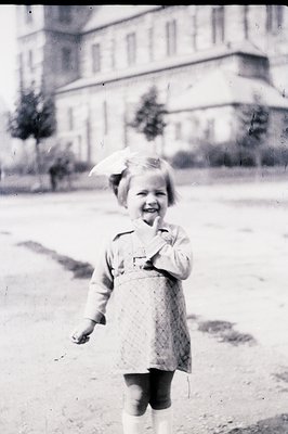 A young girl stands outdoors in a patterned dress, knee socks, and a bonnet. A large, stately building forms a blurred backdr...