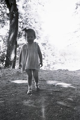 A young child in a vintage dress and sandals walks along a dirt path. The image, likely a candid snapshot, exhibits grain cha...
