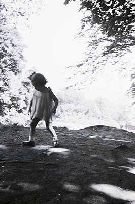 A young girl in a simple dress walks across a sloping, overgrown terrain, likely a hillside or embankment. The background is ...