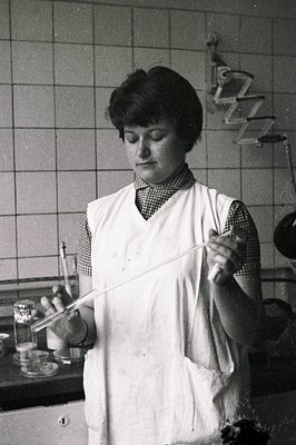 A woman in a lab apron appears to be conducting an experiment, holding a test tube. The lab background shows tile and equipme...