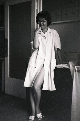 A woman in a lab coat and sandals sits on a ledge, appearing relaxed and thoughtful. The photograph, likely taken in the 1960...