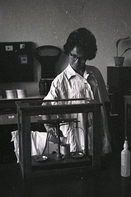 A woman in a lab coat, glasses, and short hair uses a balance scale in a laboratory setting. Equipment visible: test tubes, f...