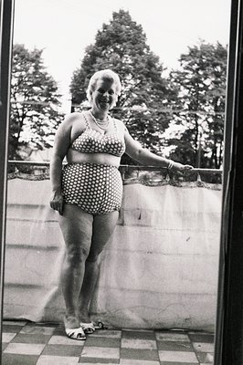 A smiling woman in a vintage, patterned two-piece swimsuit stands on a tiled balcony. She wears a pearl necklace and sandals....
