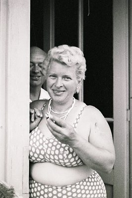 A woman in a polka dot bathing suit smiles directly at the camera. A man’s face is partially visible behind her, wearing a sh...