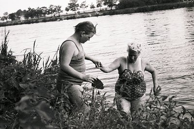 Two women wade into a calm body of water, possibly a lake or pond. They are wearing vintage swimwear, indicative of the 1940s...