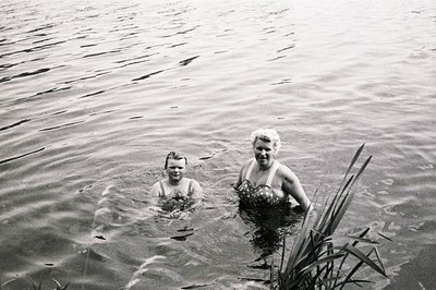 Two figures emerge from water, one youth in swim trunks and one woman in a vintage patterned swimsuit. Lush foliage is visibl...
