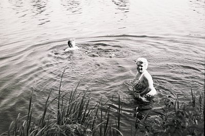 Two figures in water; one swimming, the other wading, partially obscured by tall grasses along the shoreline. High-contrast b...