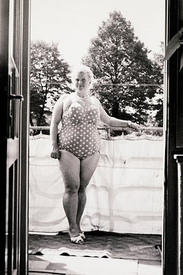 A woman in a vintage polka dot swim cap and one-piece suit stands on a balcony, captured through an open door. Architectural ...