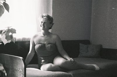 A young woman sits on a cushioned bench near a window, wearing a vintage strapless swimsuit. Indoor scene with diffused light...