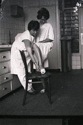 Two women in what appear to be nursing uniforms assist a seated patient, possibly in a hospital or clinic setting. Built-in c...