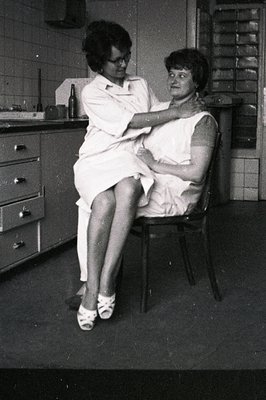 Two women in a medical setting. One, wearing a nurse's uniform and spectacles, examines the other who sits in a chair, visibl...