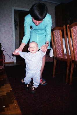 A baby stands with arms outstretched, supported by a woman in a pale blue dress. The scene appears to be a domestic interior,...