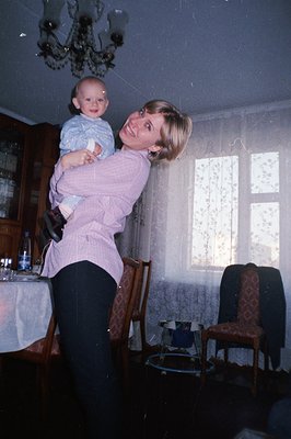 A woman in a pink button-down shirt and black trousers cradles a baby in a floral-patterned dining room. The room features a ...