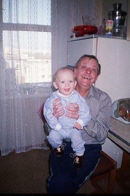 A man holds an infant, both smiling, in a kitchen setting. Visible details include lace curtains, worn wallpaper, and vintage...