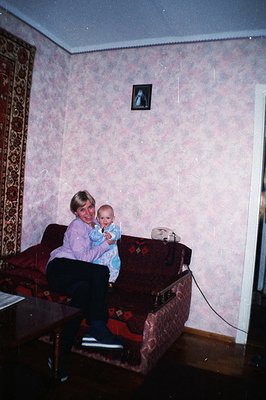 A woman holds an infant on a red velvet sofa in a room featuring patterned wallpaper and a vibrant rug. The decor suggests a ...