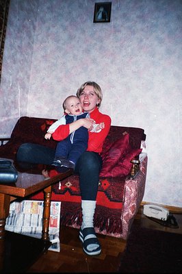 A woman in athletic wear cradles a baby in her lap while seated on a floral patterned couch. The room features wooden floors ...