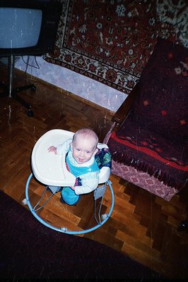 Infant sits in a blue plastic baby walker, seemingly engaged with the camera. The room features a patterned area rug and dark...