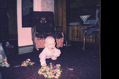 Infant crawls amidst scattered flower petals on a dark patterned floor. A richly upholstered armchair and a vintage radio cab...
